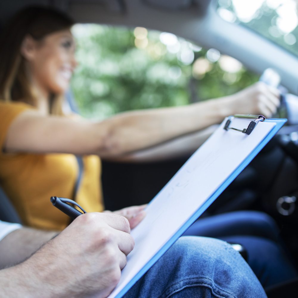 Close up view of driving instructor holding checklist while in background female student steering and driving car.