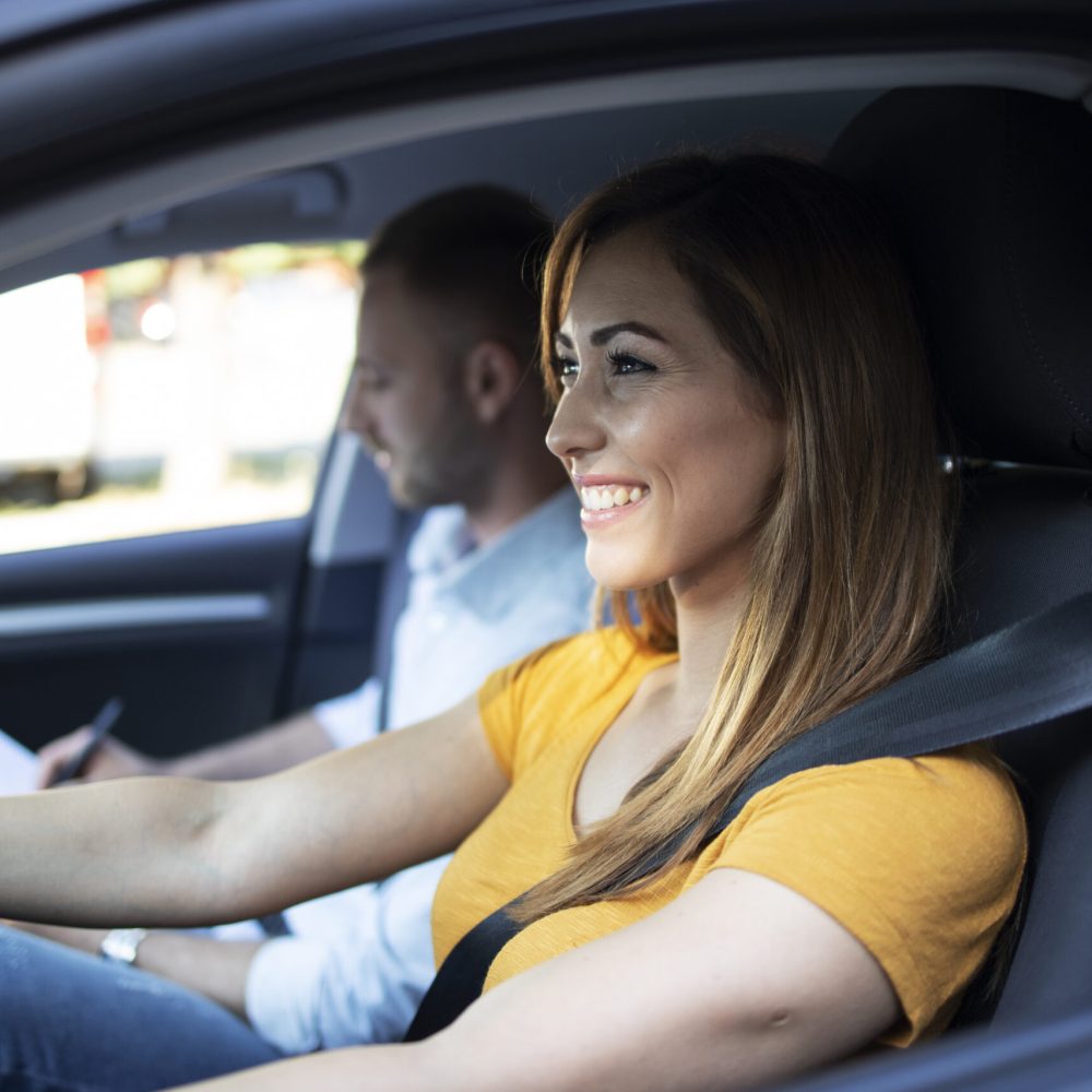 Close up view of female student driving a car and instructor holding checklist in background.